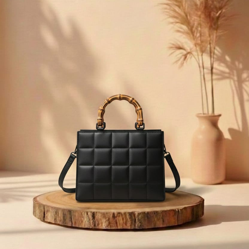 Black quilted handbag with bamboo handle on a wooden stand against a beige wall with a vase and plants.