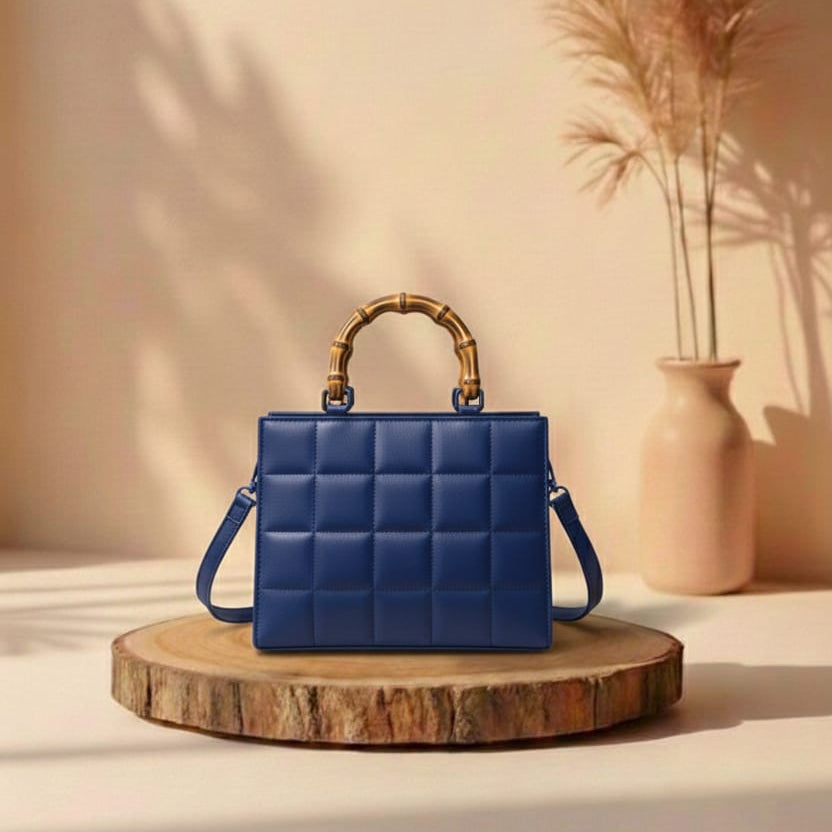 Blue quilted handbag with gold handle on a wooden stand against a beige wall with a vase of dried plants.