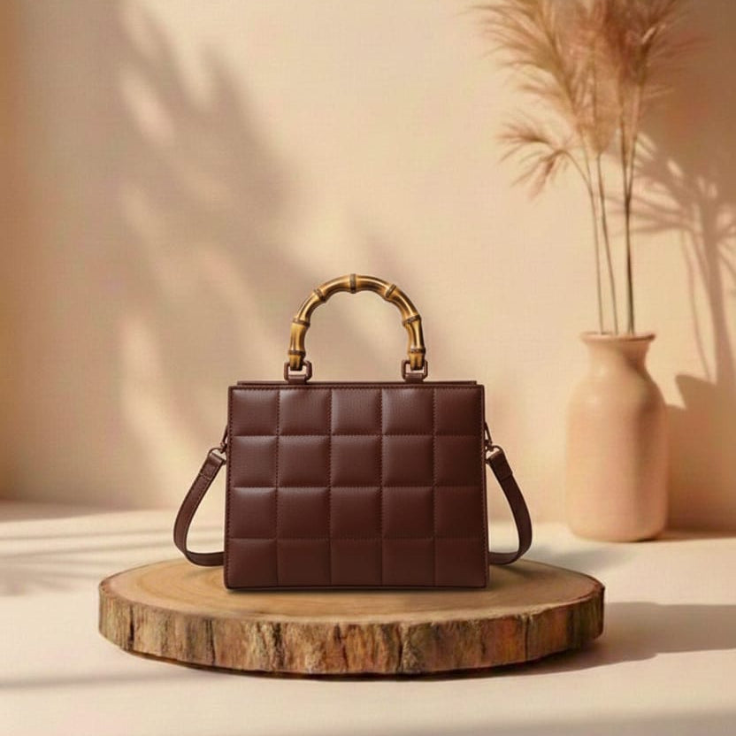 Brown quilted handbag with gold handle on a wooden platform against a beige wall with a vase of dried plants.
