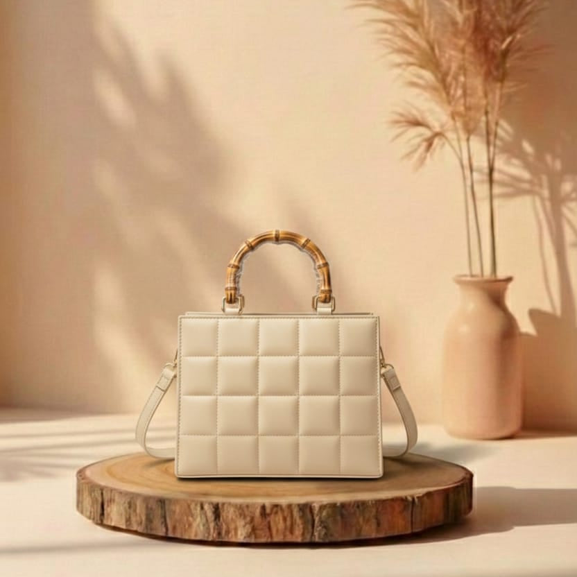 White quilted handbag with bamboo handle on a wooden stand against a beige wall with a vase and plants.