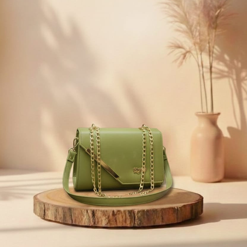 Green handbag with gold chain on a wooden platform against a beige wall with a vase and plants.