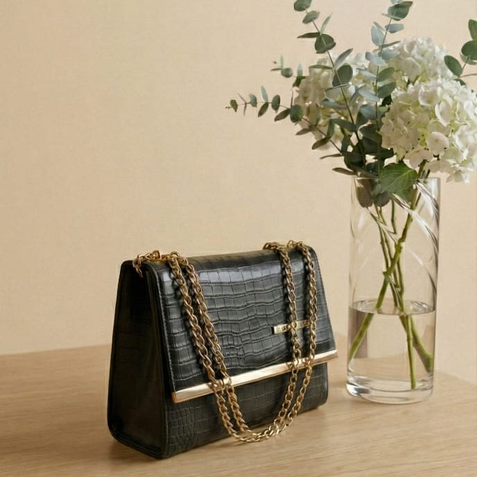 Black handbag with gold chain detail on a wooden surface next to a vase of flowers.