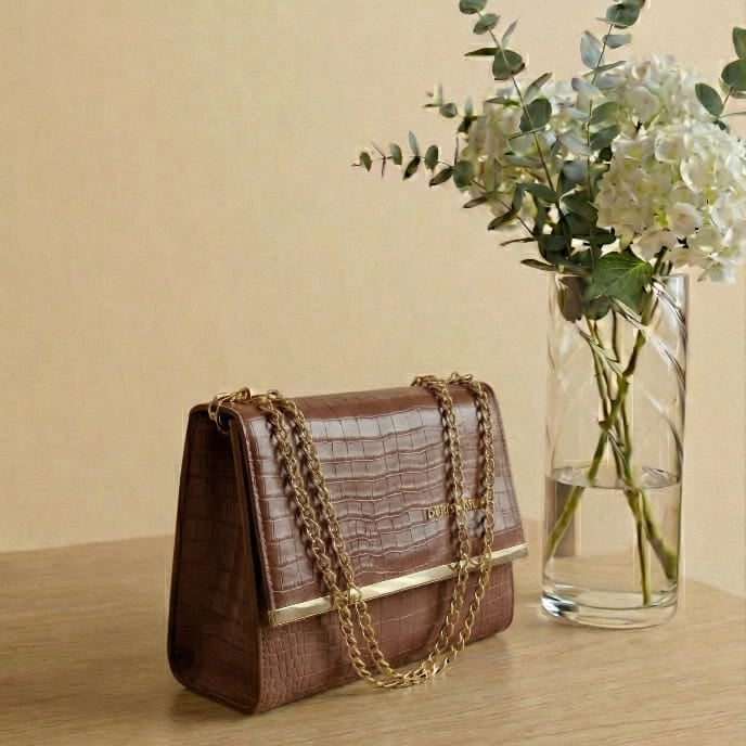 Brown handbag with gold chain on a wooden surface next to a vase with flowers.