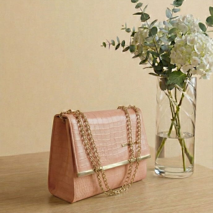 Pink handbag with gold chain on a wooden surface next to a vase with flowers.