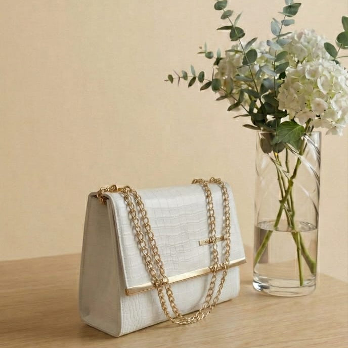 White handbag with gold chain detail on a wooden surface next to a vase of flowers.